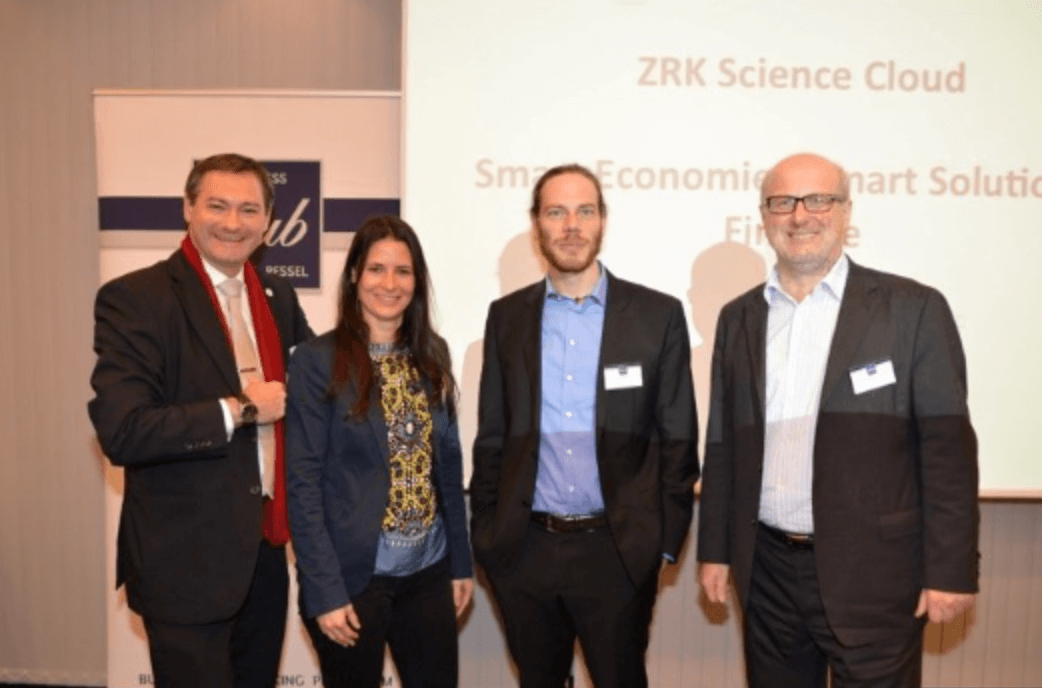 Vier Fachleute in Business-Kleidung posieren vor einer Präsentationswand mit ZRK Science Cloud und Smart-Economy-Inhalten.