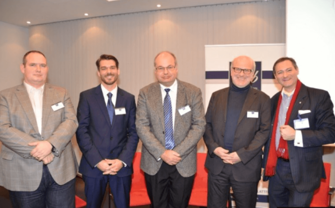 Fünf Männer in Business-Kleidung stehen nebeneinander in einem modernen Konferenzraum bei einer Veranstaltung.