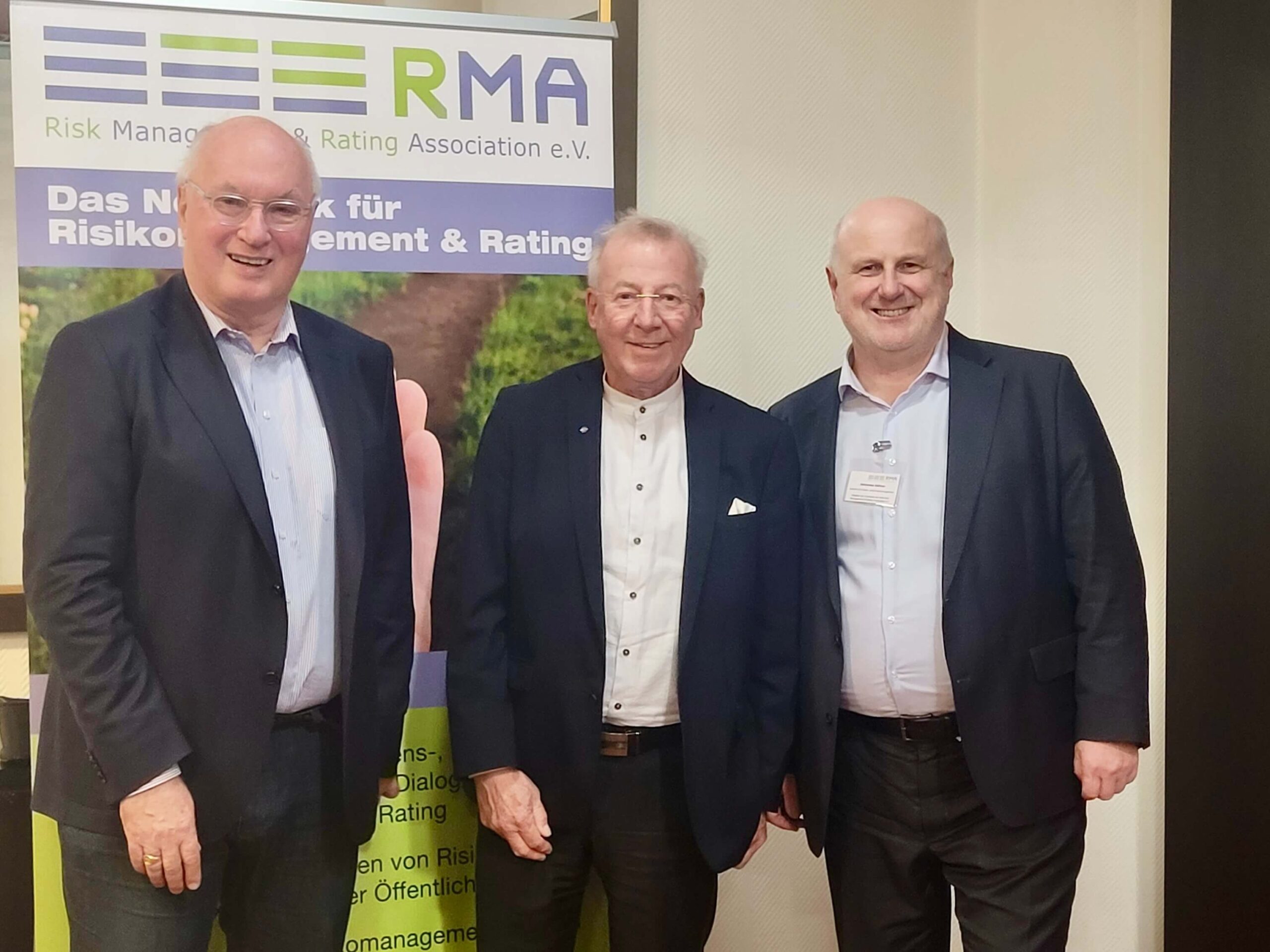 Drei Geschäftsmänner in Anzügen posieren vor dem RMA-Logoschild bei einer Veranstaltung.