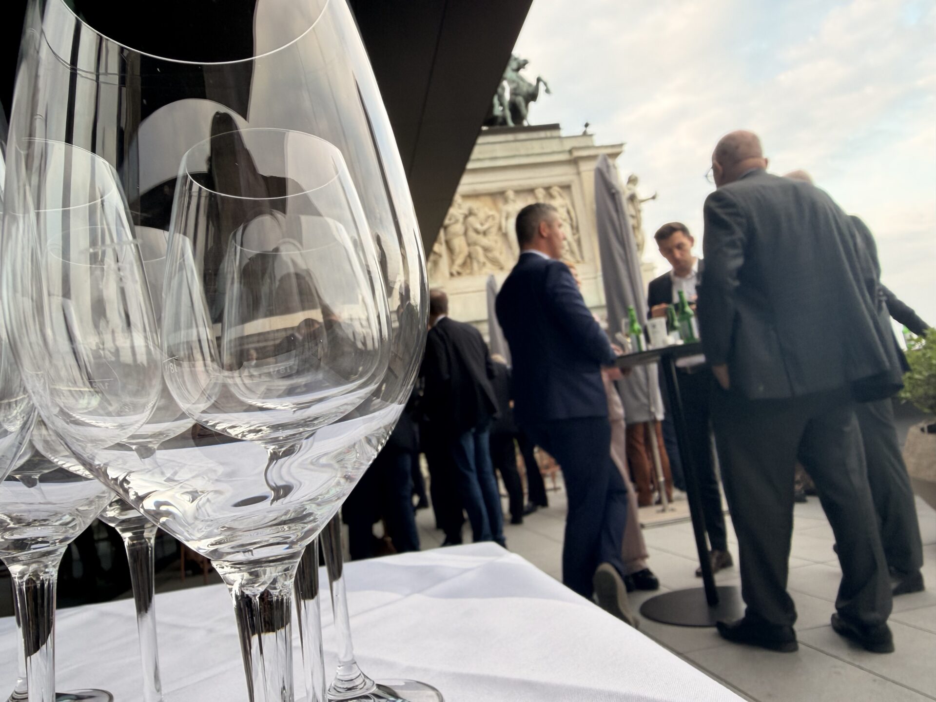 Leere Weingläser auf Tisch bei Außenevent mit Geschäftspersonen und klassischem Gebäude im Hintergrund.