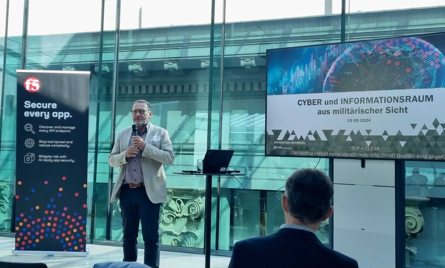 Referent spricht auf Konferenz zu Cyber und Informationsraum vor Publikum in moderner Glashalle mit Präsentationsbildschirmen.