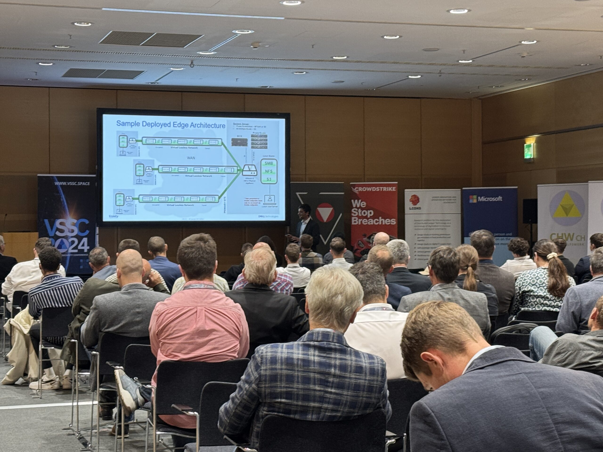 Konferenzteilnehmer verfolgen einen Vortrag zur Edge-Architektur auf Großbildschirm in modernem Konferenzsaal mit Sponsorlogos.