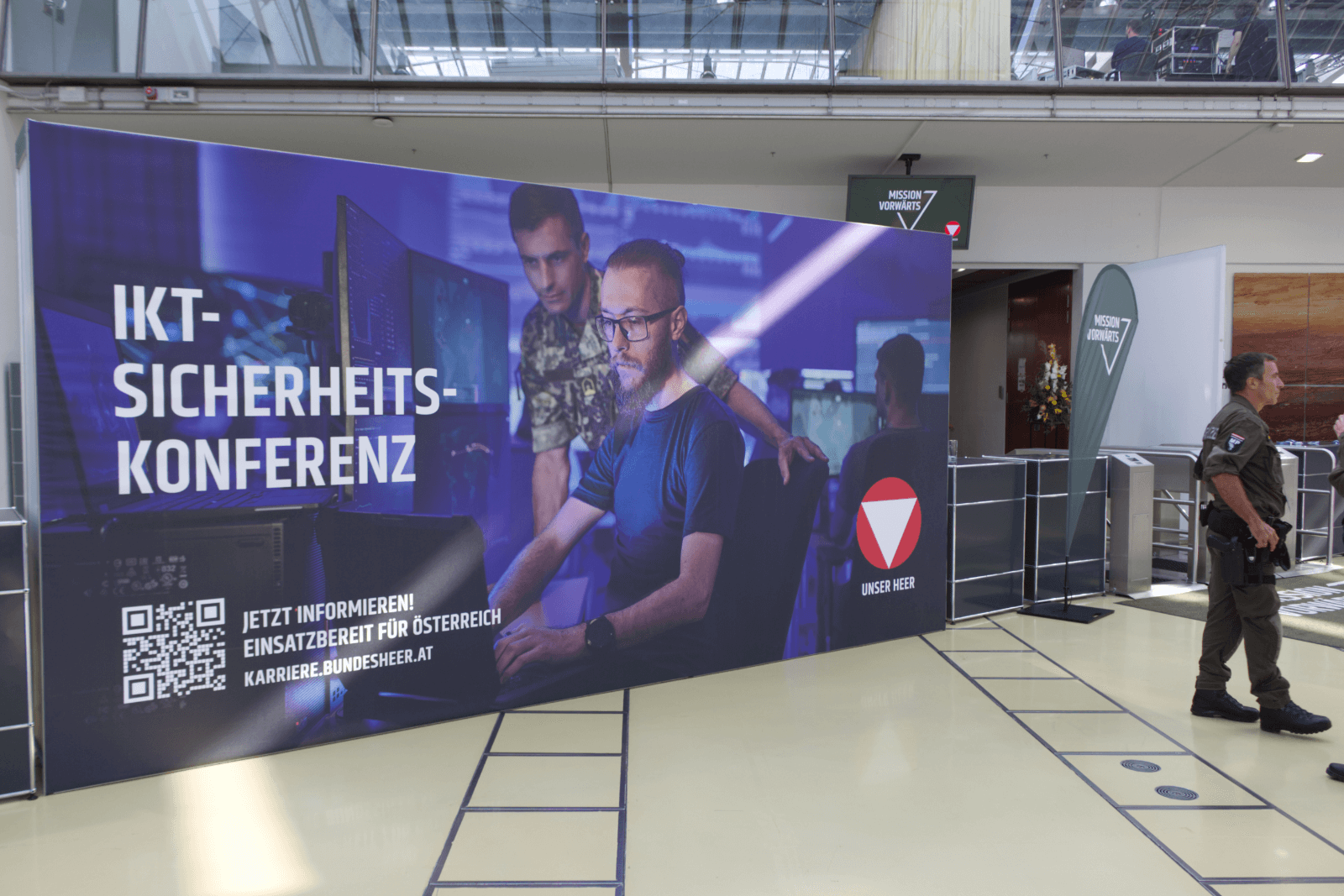 Großbildschirm mit Werbung zur IKT-Sicherheitskonferenz in einer modernen Veranstaltungshalle mit Besucher.