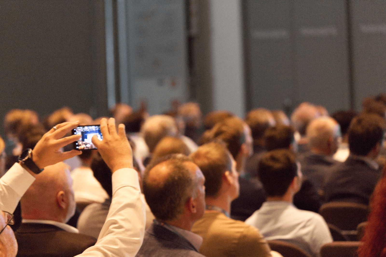 Teilnehmer einer Veranstaltung fotografiert mit Smartphone während Präsentation vor Publikum.
