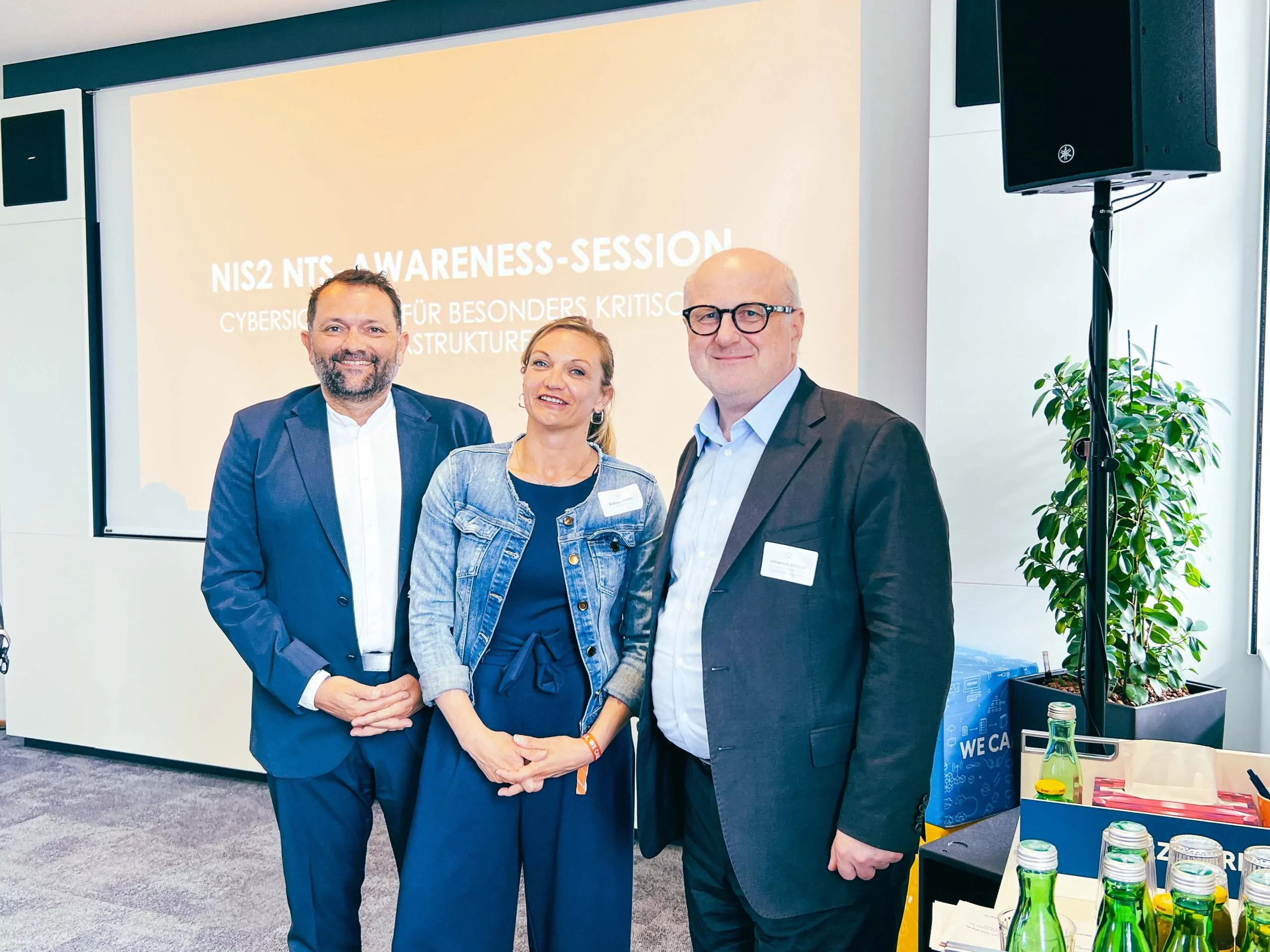 Drei Personen posieren vor einer Projektionsfläche mit NIS2-Awareness-Session-Titel in einem Konferenzraum.