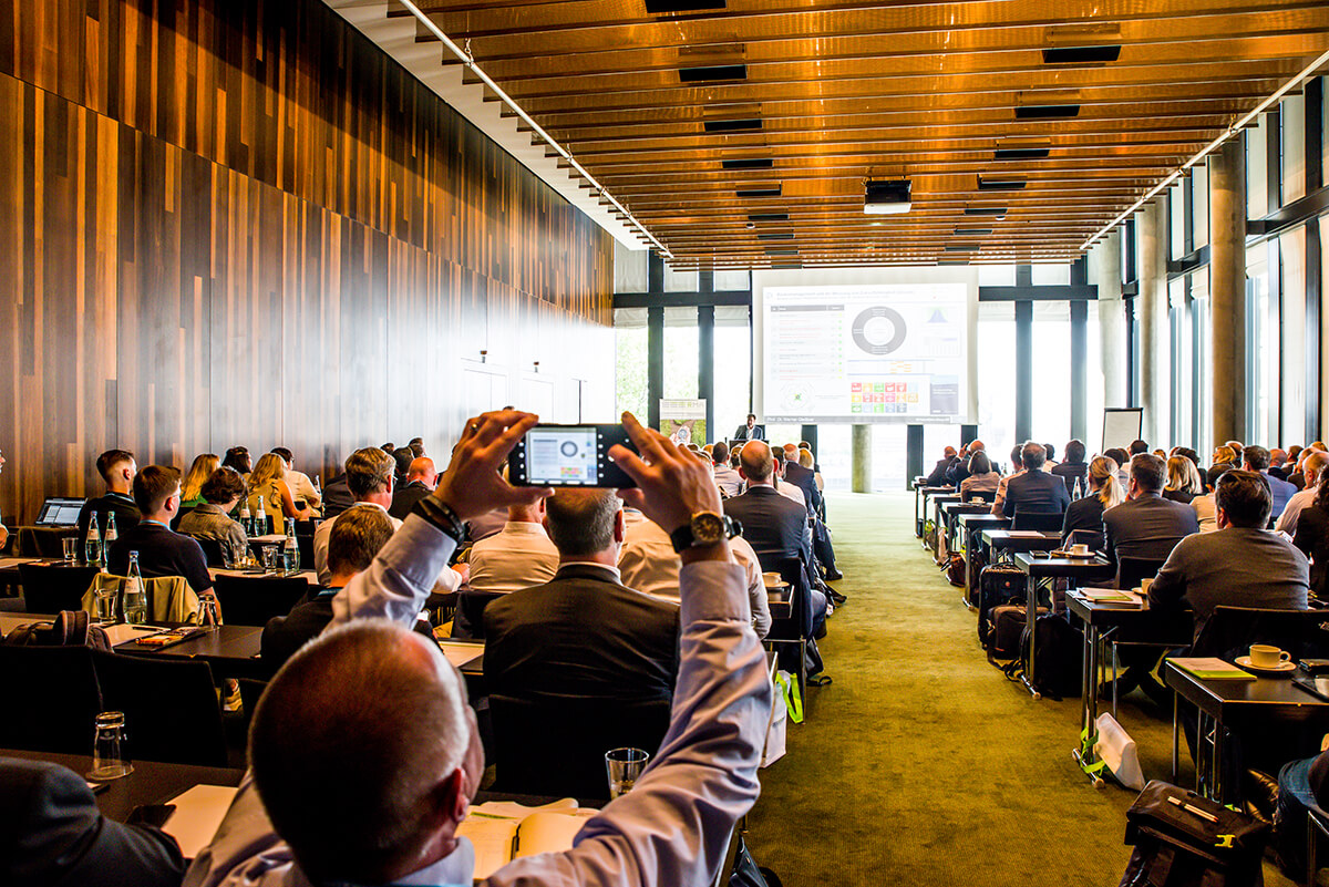Konferenzteilnehmer in großem Saal mit Holzdecke, ein Besucher fotografiert die Präsentation auf der Leinwand.