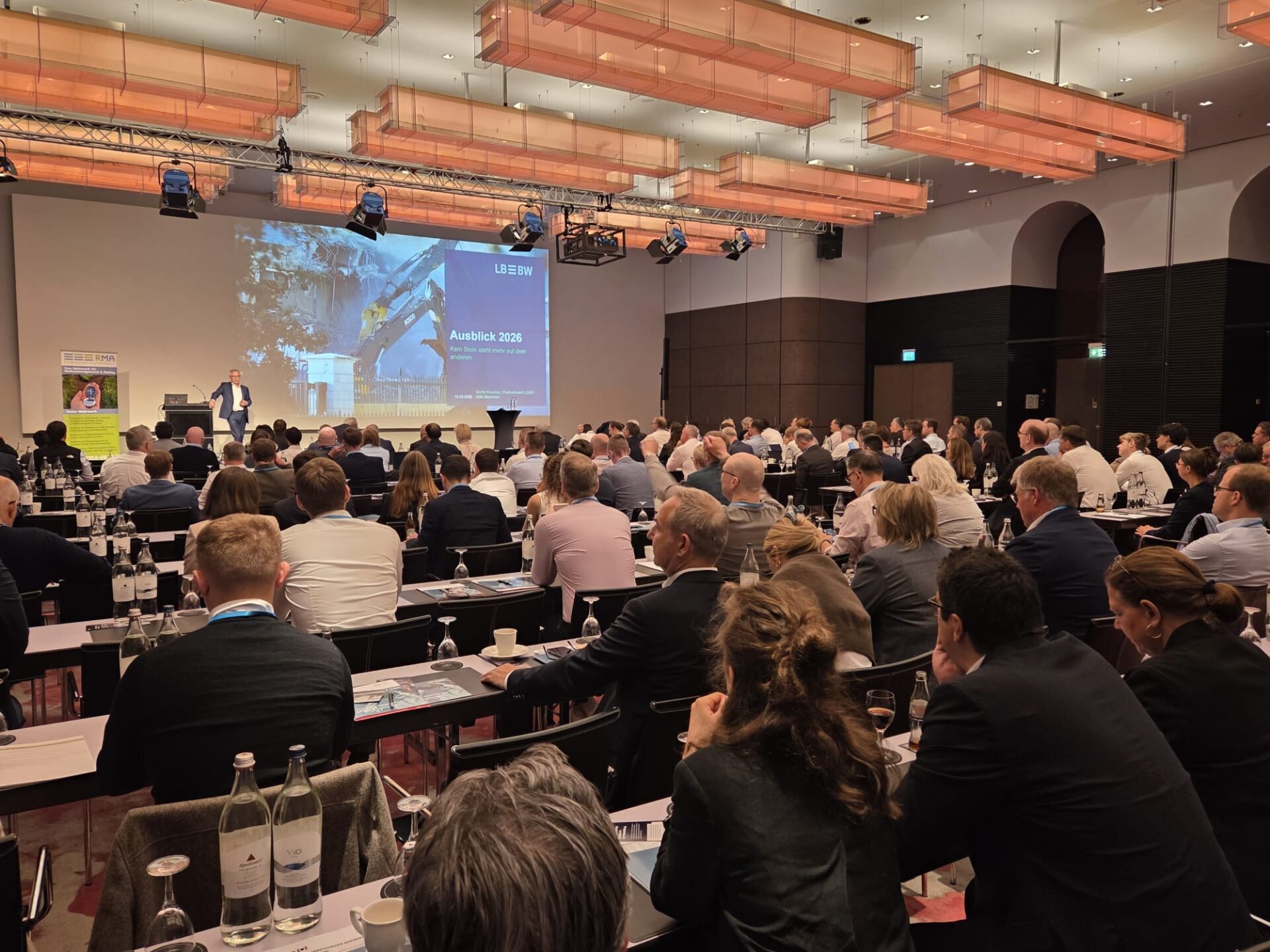 Fachkonferenz in großem Veranstaltungssaal mit Publikum an Tischen, Referent präsentiert LBBW-Vortrag "Ausblick 2026" auf Leinwand.