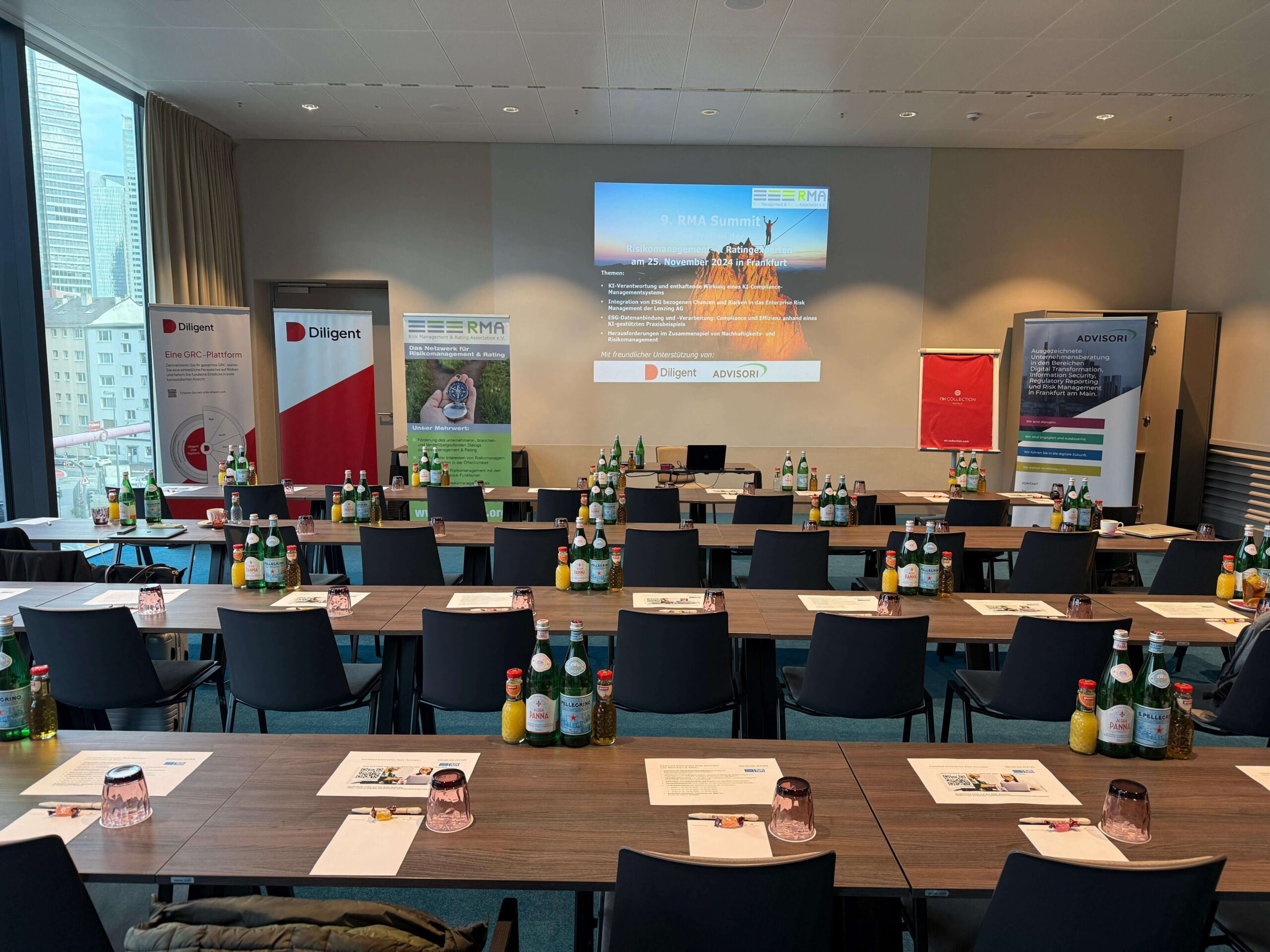 Konferenzraum mit mehreren Holztischen, schwarzen Stühlen, Getränken und Materialien, Präsentationsleinwand mit RMA-Summit-Inhalten, Diligent-Banner und Stadtblick.