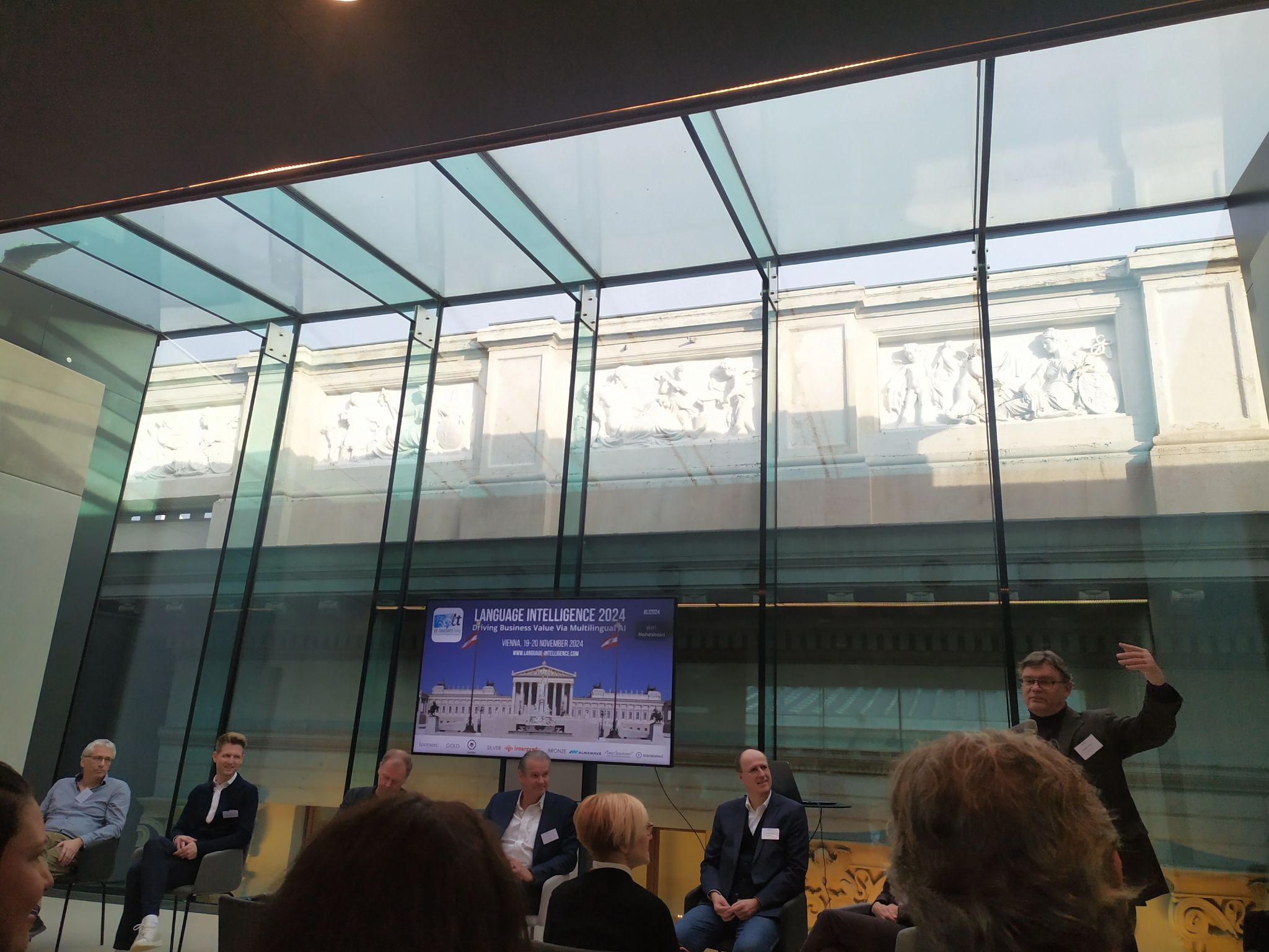 Konferenzraum mit Glasschiebewänden; Diskussionsrunde mit mehreren Teilnehmern und Präsentation zur Language Intelligence 2024 auf Bildschirm.